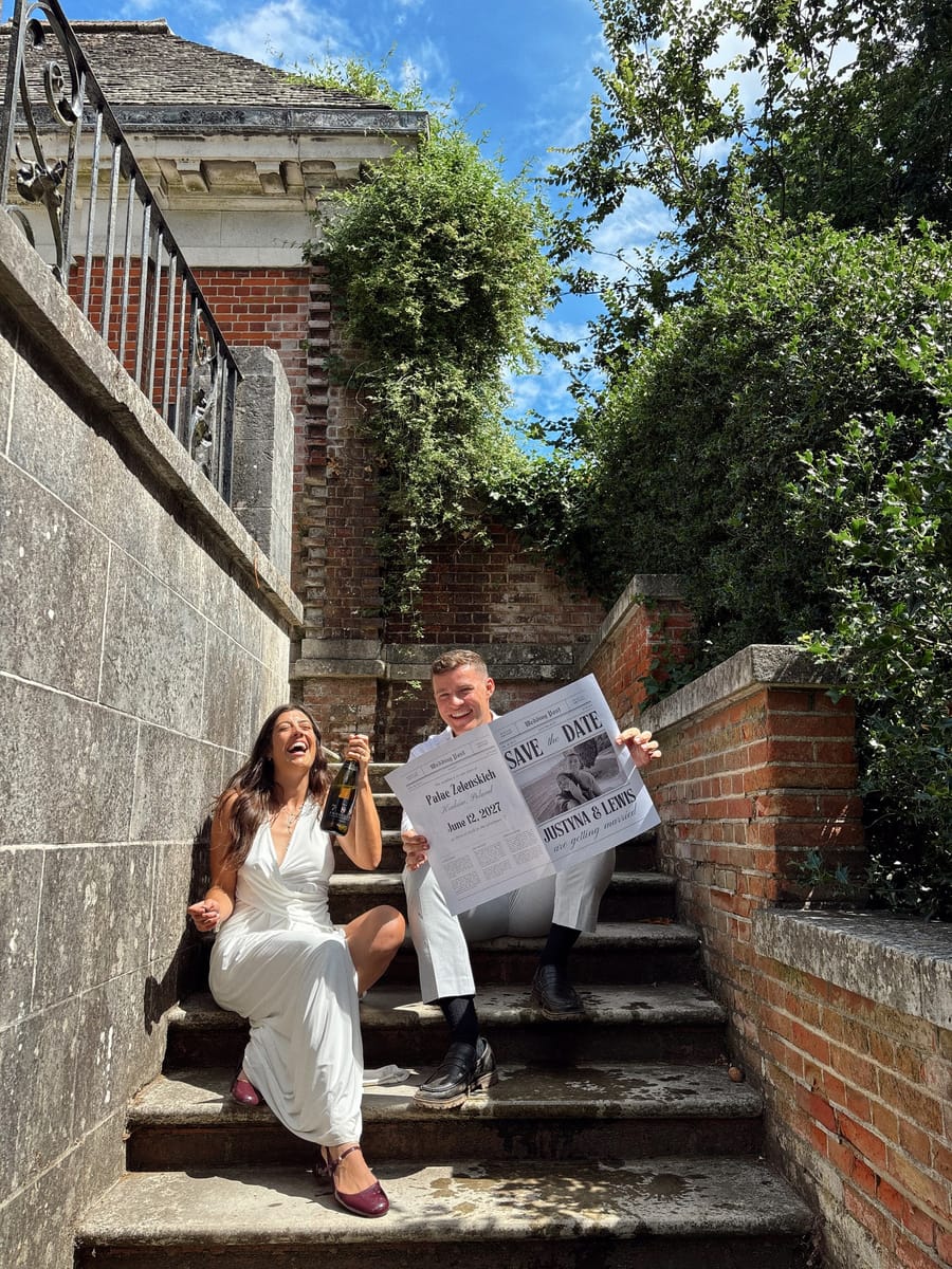 Justyna and Lewis laughing on garden steps, holding a save-the-date newspaper